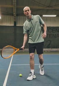 Vihreä mintunvärinen polo-paita mustilla yksityiskohdilla, mustat shortsit ja valkoiset tennarit. Pitelee oranssia tennismailaa, valmiina lyömään tennispalloa.