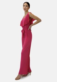 Mono fucsia con escote con volantes, textura plisada, cintura ajustada y piernas acampanadas. Un cinturón atado acentúa la cintura.