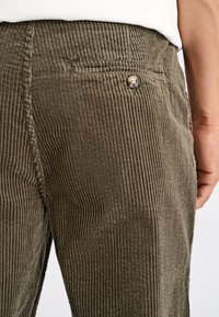 Olijfgroene corduroybroek met opvallende verticale ribbels, een standaardzak en een metalen knoopdetail aan de achterkant.