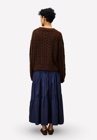 Eine Person trägt einen braunen strukturierten Pullover, einen langen, gestuften blauen Rock und braune Ballerinas, während sie mit dem Rücken zu sehen vor einem weißen Hintergrund steht.