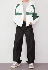 Biało-zielona kurtka wiatrówka o krótkim kroju, zestawiona z luźnymi czarnymi dżinsami i szarymi tenisówkami. Tkanina wydaje się gładka i lekka.