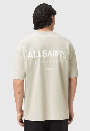 Άνδρας με σκούρα μαλλιά που φοράει μια μπεζ μπλούζα AllSaints με τα κείμενα "Στείλε μου μια καρτ ποστάλ" και "Λονδίνο" στην πλάτη, στέκεται μπροστά σε ένα απλό φόντο.