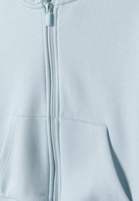 Svijetloplavi zip-up dukserica napravljena od mekane tkanine, središnjim zatvaračem i prednjim džepom s vidljivim detaljima šavova.