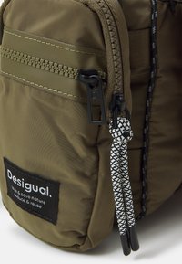 Gros plan sur un sac en tissu vert olive avec des fermetures éclair noires et des tirettes tressées blanches et noires, avec une étiquette noire affichant la marque et un message environnemental.