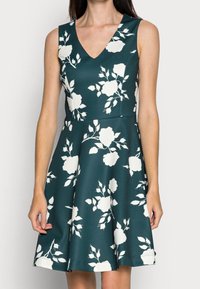 Ärmlös A-linje klänning i mörk teal med vita blommönster, med en djup V-ringning och en utsvängd kjol sydd av slät tyg.