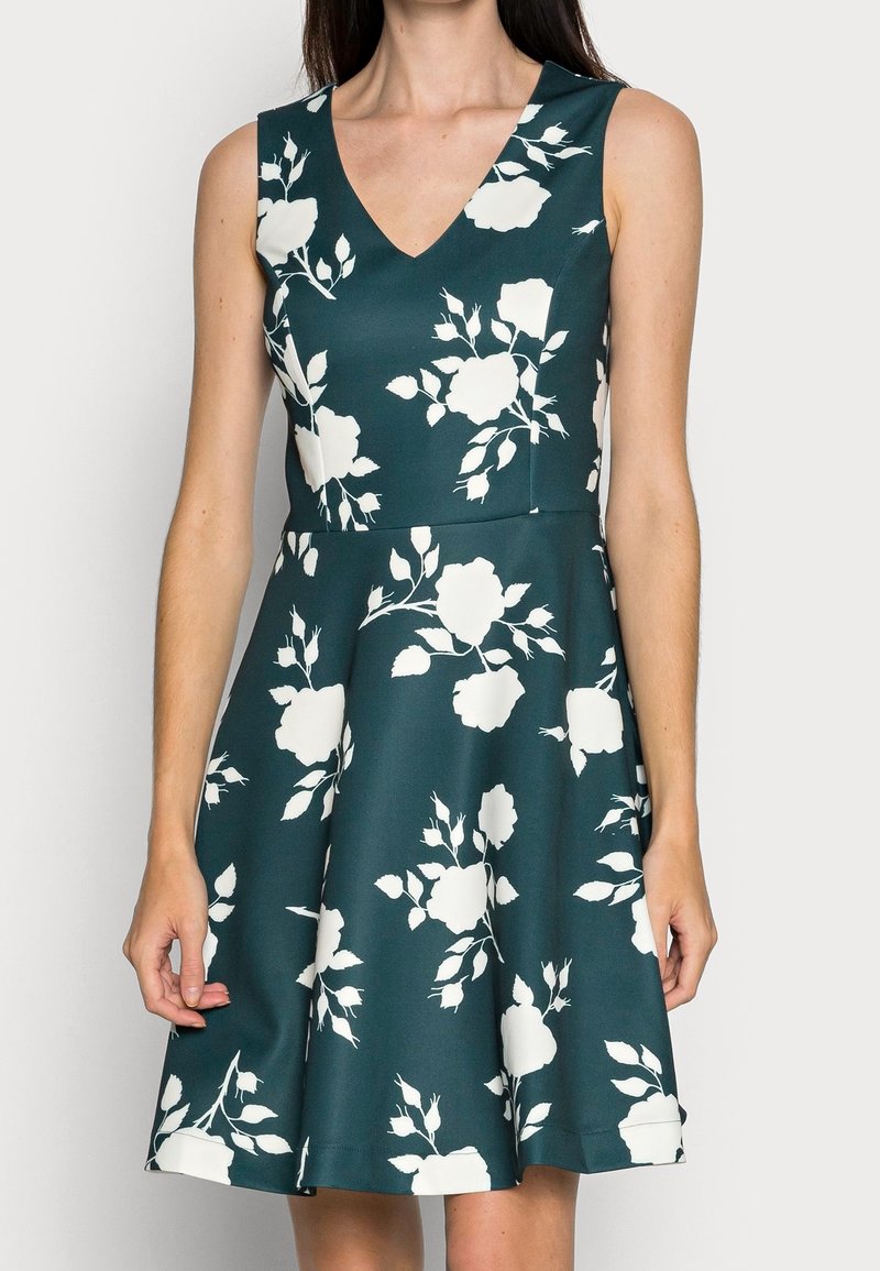 Ärmlös A-linje klänning i mörk teal med vita blommönster, med en djup V-ringning och en utsvängd kjol sydd av slät tyg.