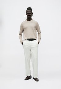 Žiemiškos smėlio spalvos megztinis ilgomis rankovėmis, šviesių plačių kelnių, juodas diržas ir rudi batai. Modelis stovi su rankomis kišenėse priešais paprastą foną.