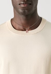 Suéter de punto color crema con un escote redondeado. Un collar de cadena plateado presenta un colgante de oso de goma rosa colocado alrededor del cuello.