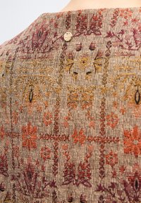 Κοντινό πλάνο υφάσματος με υφαντά γεωμετρικά και λουλουδάτα μοτίβα σε κόκκινες, πορτοκαλί και κίτρινες αποχρώσεις, με ένα μικρό χρυσό κυκλικό κουμπί.