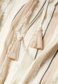 Tissu en coton à rayures horizontales beige, rose et crème. Présente des bords effilochés et deux pompons en beige clair au niveau de l'encolure.