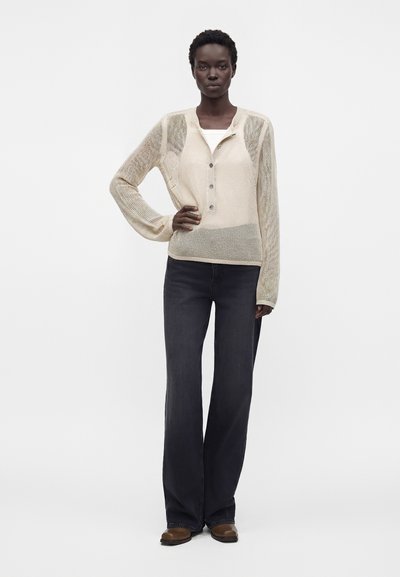 Femme debout, main sur la hanche, portant un pull en maille beige boutonné sur un haut blanc, jean noir à jambes larges et chaussures marron sur fond blanc.