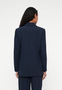 Tengerészkék, szabott blazer, strukturált szilhuettel, fokozott gallérral és hosszú ujjal. Simább anyag és egységes hátventilációs részlet.