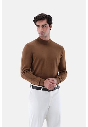 Mann mit dunklen Haaren, der einen braunen langärmligen Rollkragenpullover und weiße Hosen mit einem schwarzen Gürtel trägt, steht vor einem weißen Hintergrund.