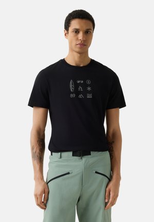 Man draagt zwart T-shirt met kleine grijze iconen gerangschikt in drie rijen, gecombineerd met lichtgroene broek met zwarte zakken met rits.