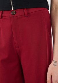 Red tailored pants with a smooth texture, featuring side pockets and a contrasting button detail at the waist.