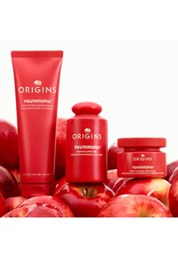 Kolme punasta ihonhoitotuotetta on esillä: putkilo, purkki ja pyöreä pullo, kaikki nimeltään "YOUTHTOPIA", ympäröityinä punaisilla omenoilla.