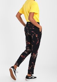 Svarta byxor med ett blommigt paisleymönster, med en figursydd passform och elastisk midja, kombinerade med en gul t-shirt och svarta sneakers.