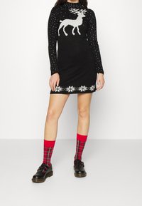 Svart stickad klänning med vitt rendesign och snöflinge-slagskärning, med långa prickiga ärmar. Matchad med röda rutiga strumpor och svarta skor.