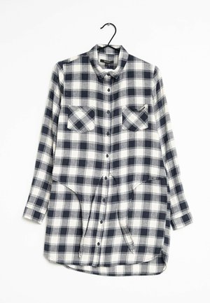 Chemise à manches longues à carreaux noirs et blancs avec deux poches poitrine et un col, suspendue sur un cintre noir.