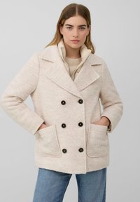 Junge Frau in einem beigen, doppelt geknöpften Wollmantel mit schwarzen Knöpfen und blauen Jeans, die mit den Händen in den Manteltaschen steht.