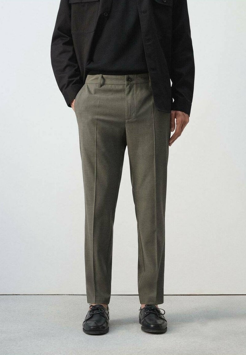 Homme portant un pantalon vert olive sur mesure, une chemise noire, une veste noire et des chaussures noires à lacets, debout devant un fond clair uni.