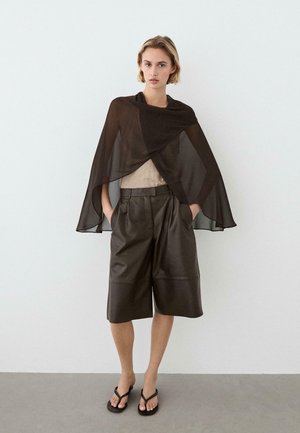 Kobieta stoi przy białej ścianie, ubrana w przezroczystą czarną pelerynę, beżowy top, szerokie ciemnobrązowe spodenki do kolan i czarne klapki, z rękami w kieszeniach.