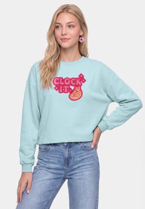 Kvinde med langt blondt hår iført lyseblå sweatshirt med pink tekst "CLOCK IT" og håndgrafik, kombineret med blå jeans og pink øreringe.