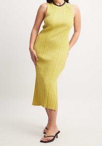 Robe jaune sans manches en tissu côtelé, silhouette ajustée, longueur au genou, fente latérale. Portée avec des sandales noires à lanières.