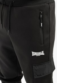 Melnas sporta bikses, kas izgatavotas no mīksta auduma, ar kabatu ar rāvējslēdzēju, Lonsdale logotipa uzlīmi un struktūru sānu panelī. Iekļauta elastīga josta.