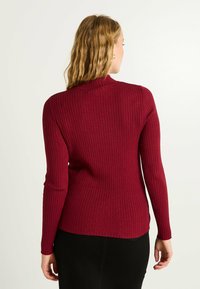 Pull en rib à col haut, de couleur cramoisie, avec des manches longues et une coupe ajustée. Présente une texture subtile et un ourlet légèrement incurvé.