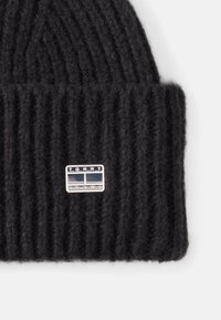 Black knitted beanie with a ribbed texture, featuring a rectangular silver logo patch with “TOMMY JEANS” in black letters.
