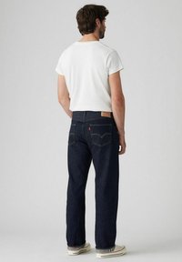 Jeans de mezclilla azul oscuro con corte recto, que presentan bolsillos traseros y un parche de cuero con el logo en la cinturilla. Combinados con una camiseta blanca.