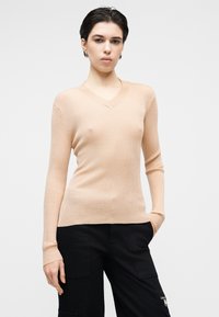 Pull à manches longues côtelé beige clair avec col en V, présentant une coupe ajustée et une texture douce. Associé à un pantalon cargo noir.