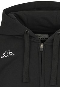 Zwarte zip-up hoodie van zachte stof, met een kangoeroezak, trekkoorden en een klein wit Kappa-logo op de linkerkant van de borst.