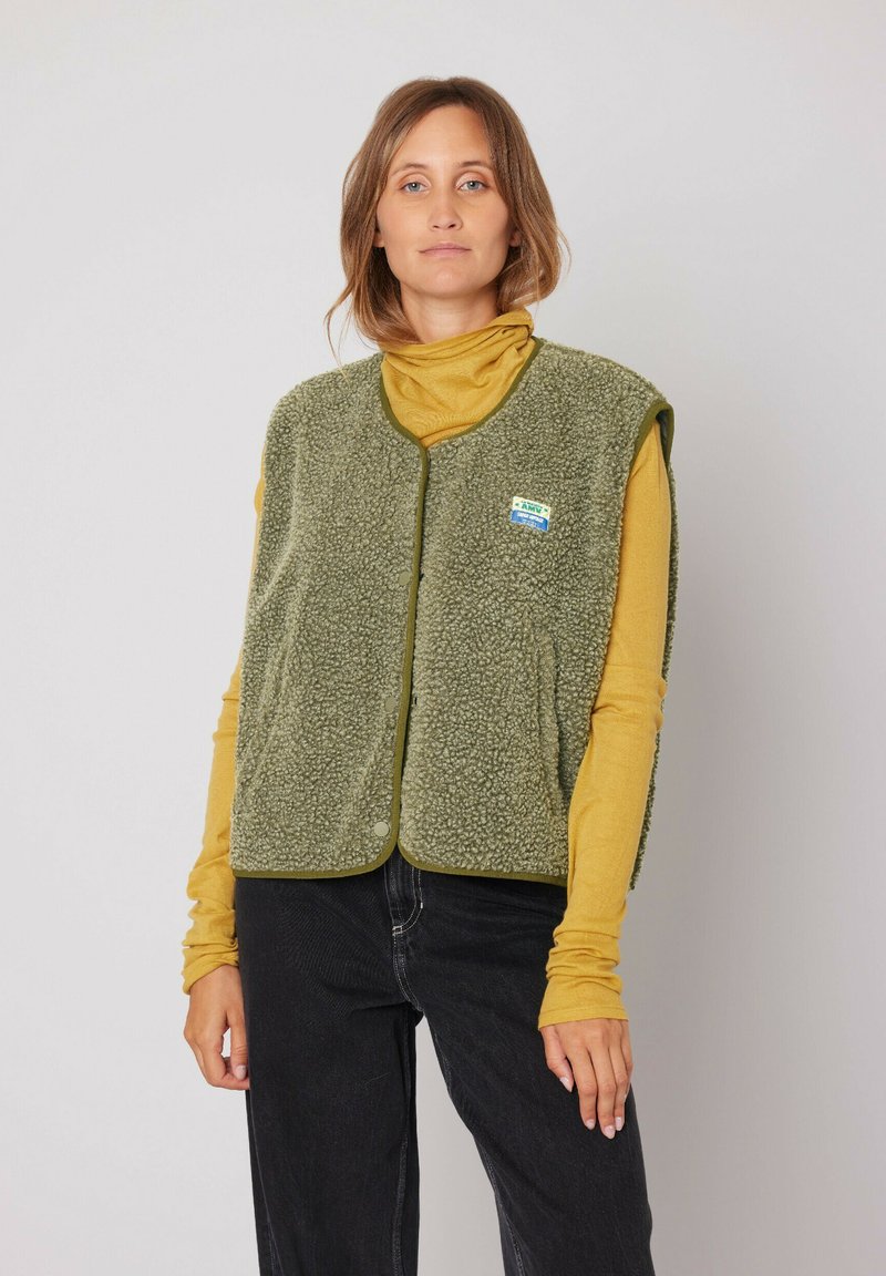 Fleece vest in olijfgroen met een zachte textuur, ronde halslijn en drukknoopsluiting. Gekocht over een mosterdkleurige longsleeve, voorzien van merkpatch.