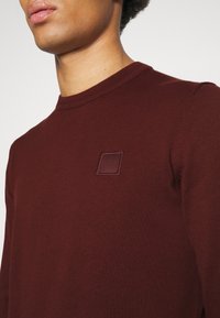 Bordeauxfarbenes Strickpullover mit Rundhalsausschnitt. Verfügt über eine strukturierte Oberfläche und ein quadratisches Patch mit dem Markennamen "BOSS" auf der Brust.