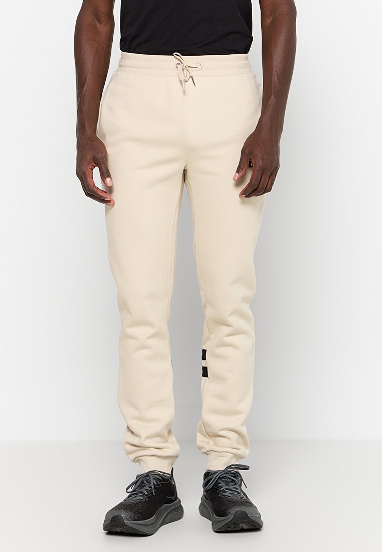 Hombre vistiendo pantalones jogger beige con cintura elástica y zapatillas negras, de pie frente a un fondo blanco liso.