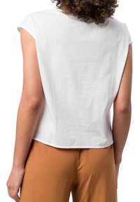 Top blanco de manga corta hecho de una tela ligera, con un corte relajado y dobladillo redondeado, combinado con pantalones marrones.