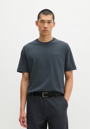 Mann trägt ein schlichtes dunkelblaues T-Shirt und passende Hose mit einem schwarzen Gürtel, steht vor einem schlichten hellen Hintergrund.