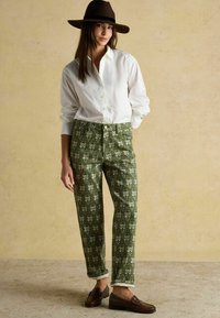 Zelené nohavice so vzorom kvetov, skombinované s bielou košeľou na gombíky a hnedými mokasínami. Modelka má na hlave hnedý klobúk a ruky opreté o boky.