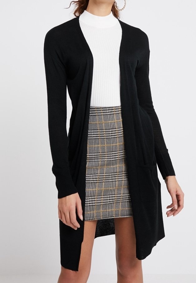 Svart knälång cardigan med öppen front, långa ärmar och fickor, lager på lager över en vit ribbad topp och en rutmönstrad kjol. Slät stickad tyg.