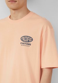 Perzikkleurig katoenen t-shirt met een donkerblauw ovaal logo en de tekst "PACIFIC CUSTOMS BOARDS&KITS" op de linkerkant van de borst.