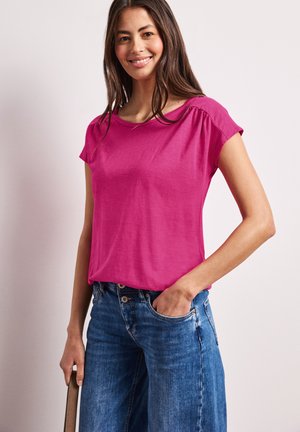 Kobieta uśmiechająca się, ubrana w jaskraworóżowy top z krótkim rękawem i niebieskie dżinsy, stoi z jedną ręką w kieszeni, a drugą trzyma brązowy pasek.