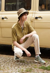 Khaki short-sleeve shirt with two chest pockets, cream pants, and green sneakers; wearing a beige bucket hat with a black accent and drawstring.