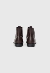 Botas de tobillo de cuero marrón oscuro con una textura suave, que cuentan con una suela negra baja y una pestaña en la parte posterior para facilitar su uso.