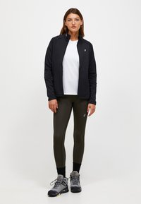Giacca nera con zip su una camicia bianca, abbinata a leggings verde oliva e scarpe da trekking grigie. Realizzata in un tessuto morbido e con un design aderente.