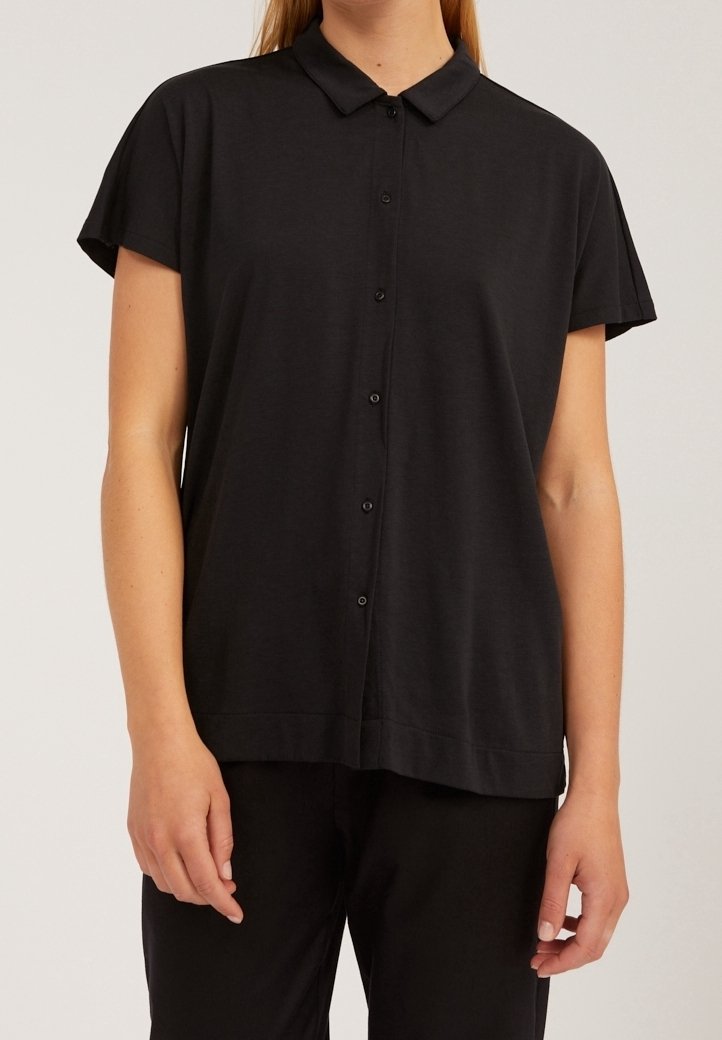 Femme portant une chemise noire à manches courtes avec col et un pantalon noir sur un fond uni.