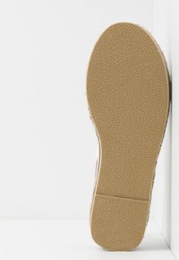 Semelle d'une chaussure beige avec une semelle extérieure en caoutchouc texturée, présentée à la verticale contre une surface d'angle blanche.
