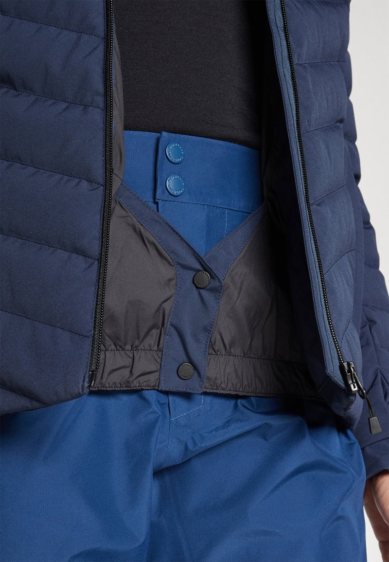 Padded navy Blouson, offen mit einem blauen Taillenbund, der Druckknöpfe aufweist und eine glatte Textur hat; geschichtetes Design mit kontrastierenden Materialien.