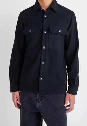 Musta nappipaita, joka on valmistettu tekstuurista kankaasta. Paita sisältää kauluksen, kaksi rintataskua ja vastaavat napit.